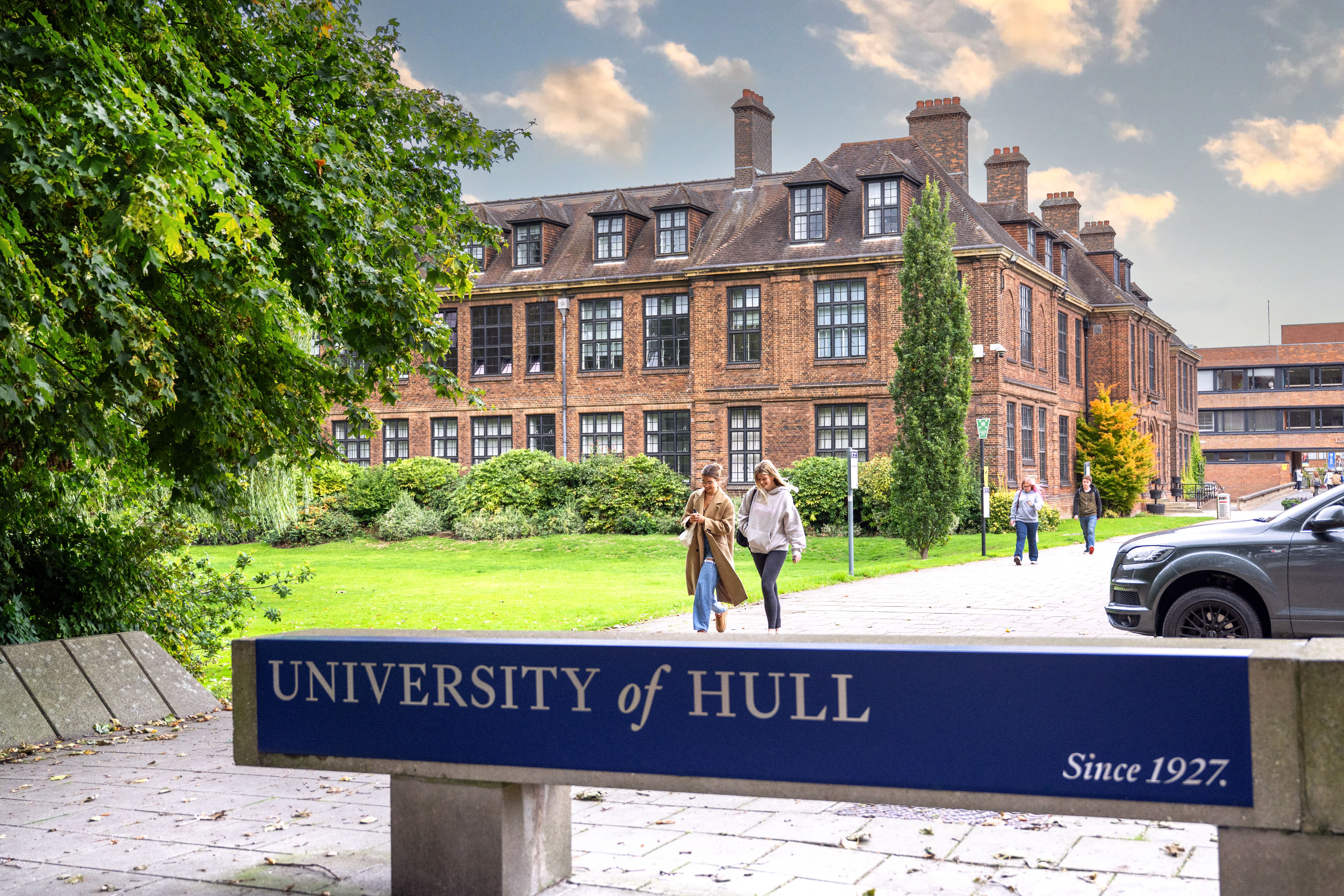 The University of Hull sign with the campus in the background and students walking