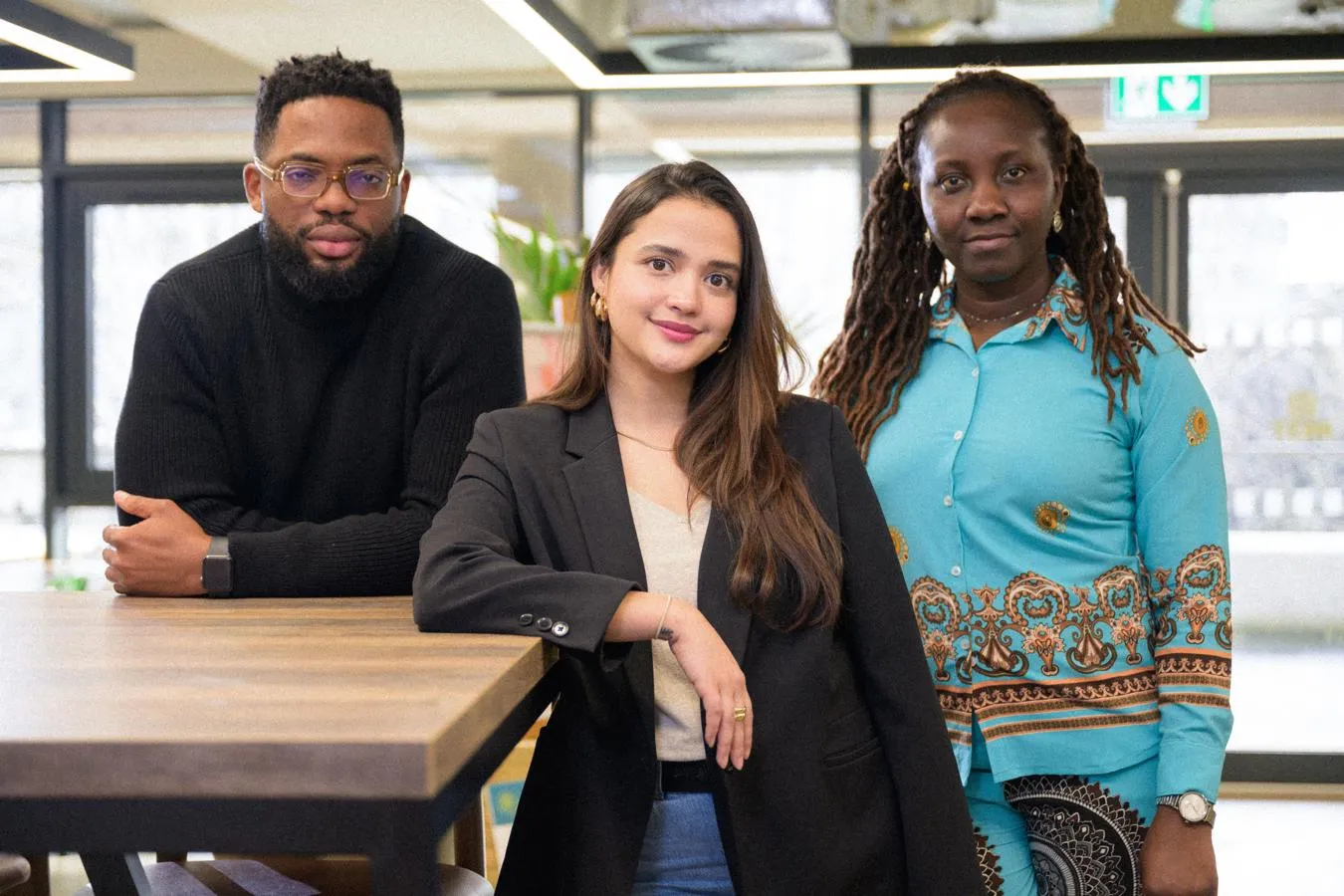 three international students standing in front of windows, two leaning against a high desk and the third at their side.