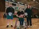 three people in a sports hall two standing either side of a man in a wheelchair, all holding balls