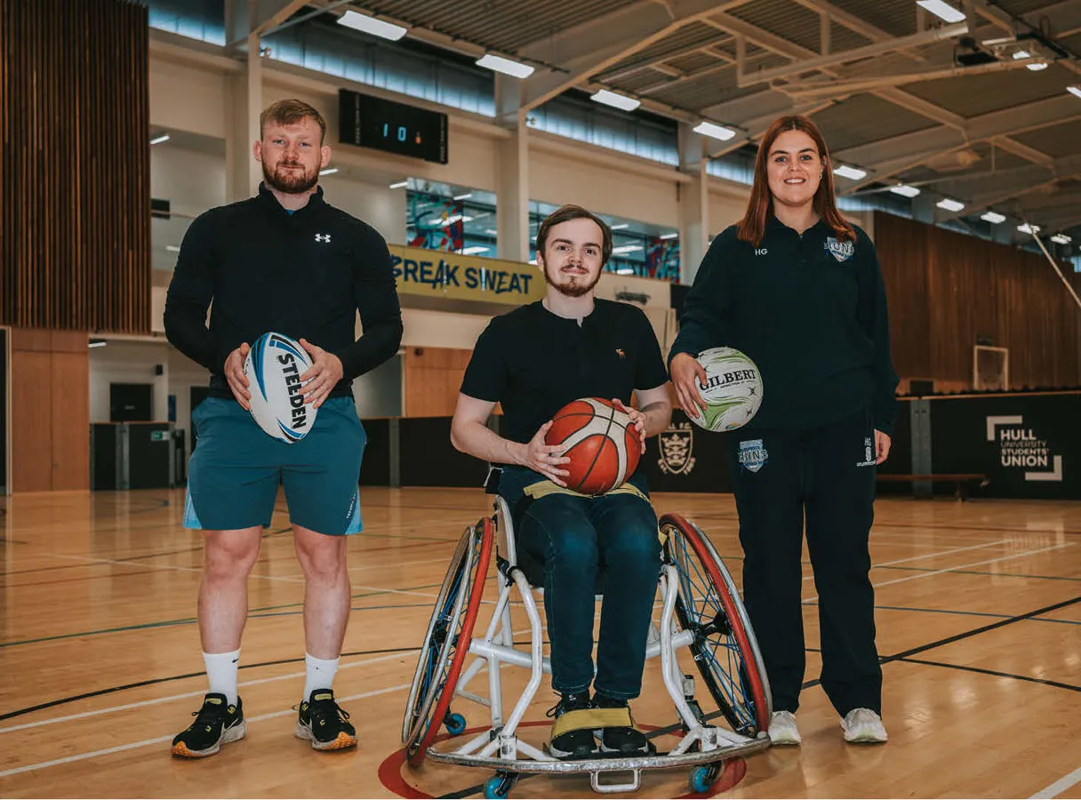 three people in a sports hall two standing either side of a man in a wheelchair, all holding balls