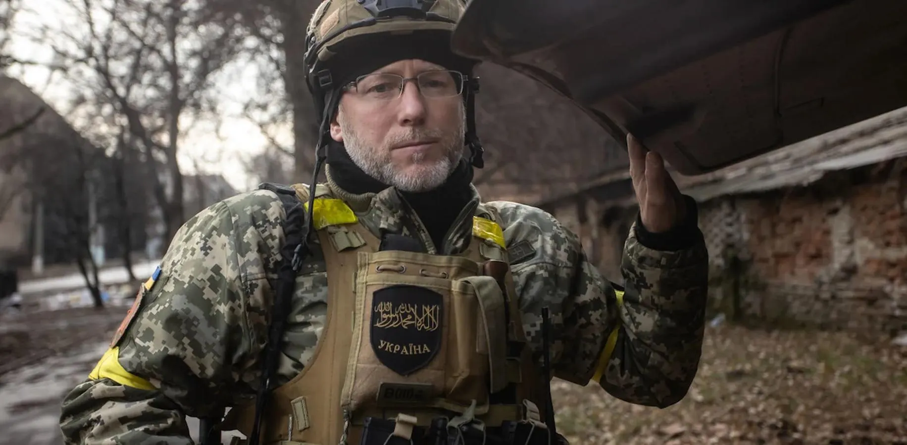 grey haired bespectacled man in camouflage and body armour pictured by a wall in a wintery urban scene