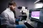 A researcher in white lab coat examines a microscopic image on a computer screen