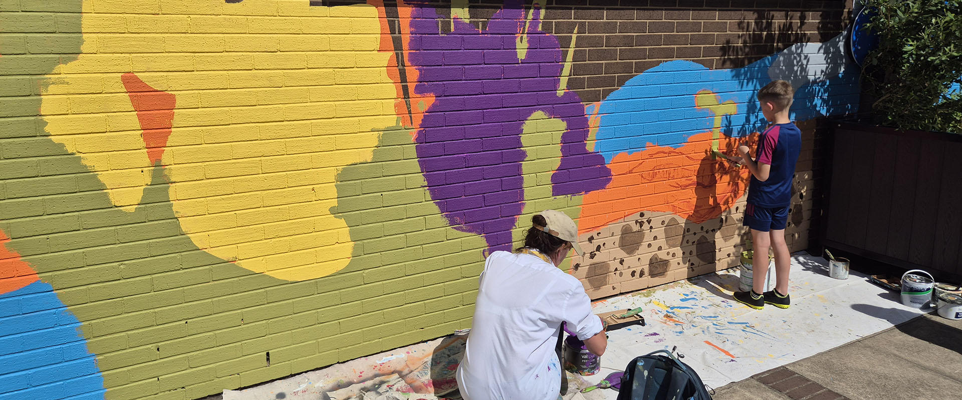 An artist and school pupil paint part of a colourful mural on a school wall
