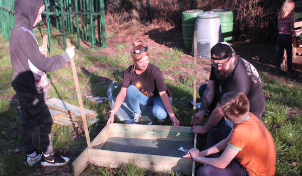 A group of children are outdoors with two youth workers, creating a composting box out of wood